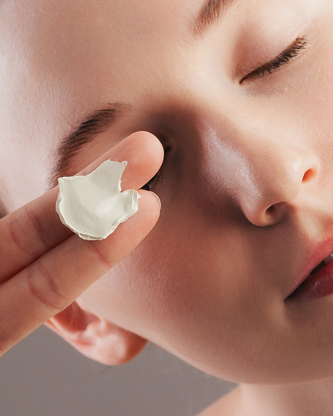 Cream on two fingers is held in front of woman's face